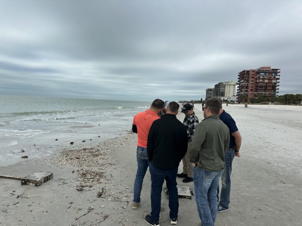 Six men stand on a Gulf beach looking at concrete slabs and footings exposed in the sand, with condominium towers along the dune line behind them. Shoreline and seawall project site walk by Anderson Dock and Lift, Sarasota County FL.