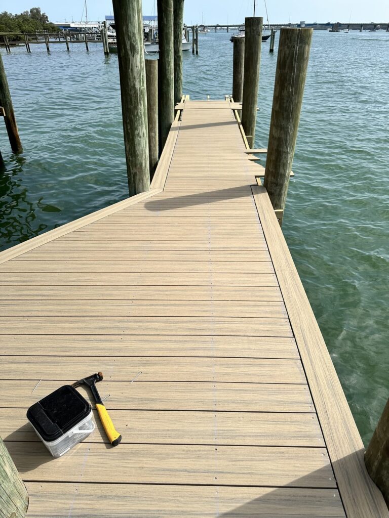A dock walkway decked in composite boards runs over the water on wood pilings, with a hammer, loose nails, and a fastener tub on the fresh decking. Dock construction by Anderson Dock and Lift, Sarasota FL.