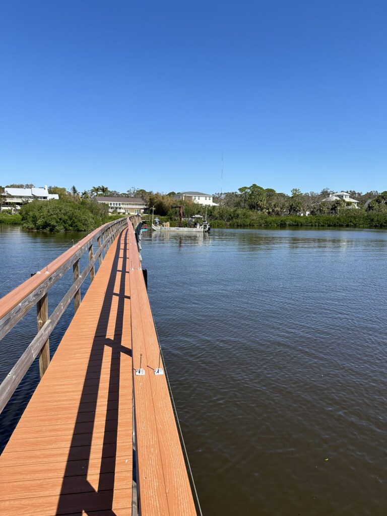 Long dock walkway with wide tan deck boards running out over calm water to a dock at the far end. Dock and pier construction by Anderson Dock and Lift, Manatee County FL.
