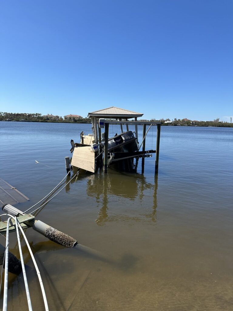 A wood dock with a peaked roof over the boat slip holds an outboard boat on a lift at low tide, with barnacle covered pilings in the foreground. Boat lift and dock construction by Anderson Dock and Lift, Bradenton FL.