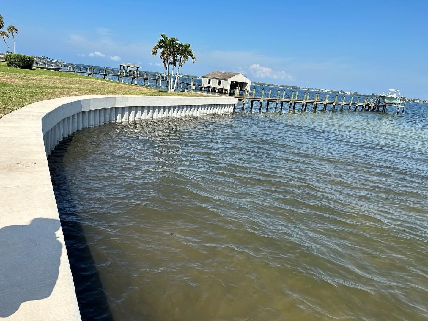 Curved concrete seawall with a new cap along a bayfront lawn, with a fishing pier and boathouse behind. Seawall construction, Anderson Dock and Lift, Manatee County FL.