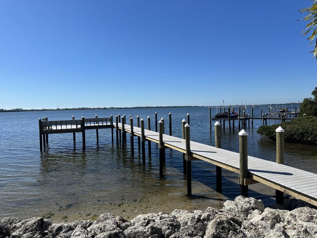 A dock walkway on capped wood pilings runs from a rock shoreline out to an L shaped platform over the bay, seen from the side. Dock and pier construction by Anderson Dock and Lift, St. Petersburg FL.