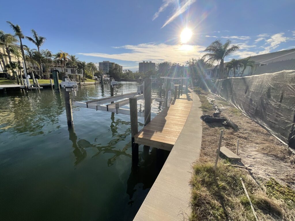 A wood plank walkway runs along a concrete seawall cap beside an aluminum boat lift standing in a canal. Dock and boat lift installation by Anderson Dock and Lift, Fort Myers FL.