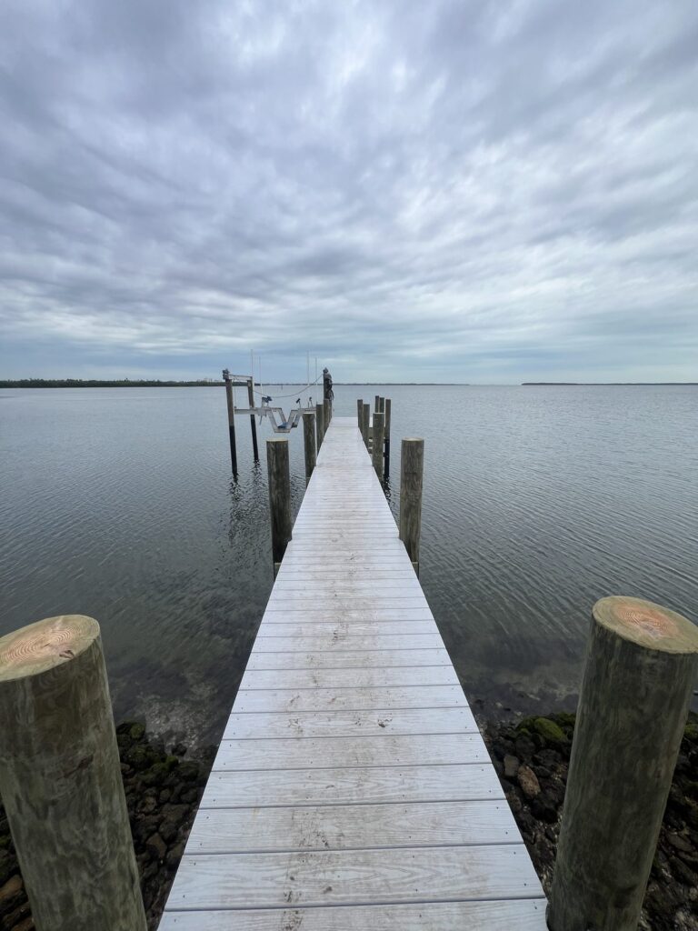 Dock walkway of light gray deck boards running out over riprap rock to open water, flanked by round wooden pilings. Dock and pier construction by Anderson Dock and Lift, Palmetto FL.