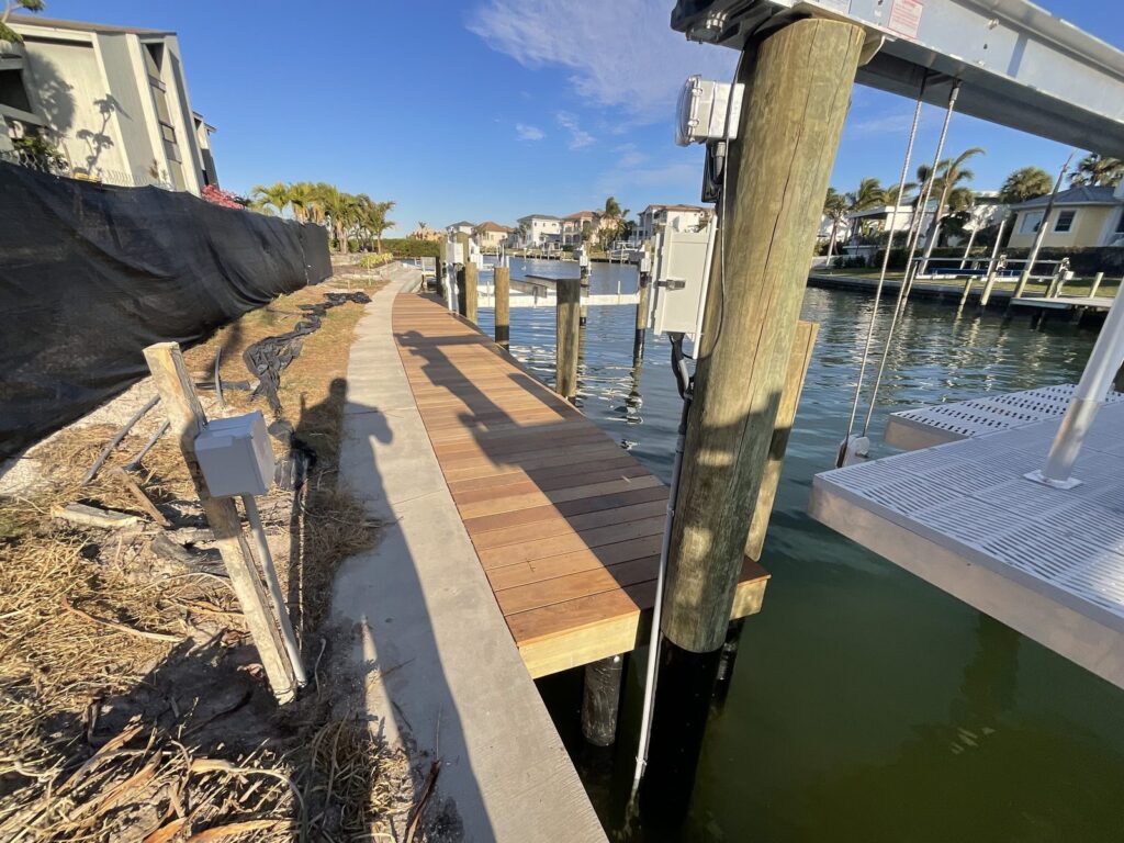 A wood plank walkway and concrete seawall cap run past a piling with mounted electrical boxes, with the grated deck of a boat lift at right. Dock and boat lift installation by Anderson Dock and Lift, Palmetto FL.
