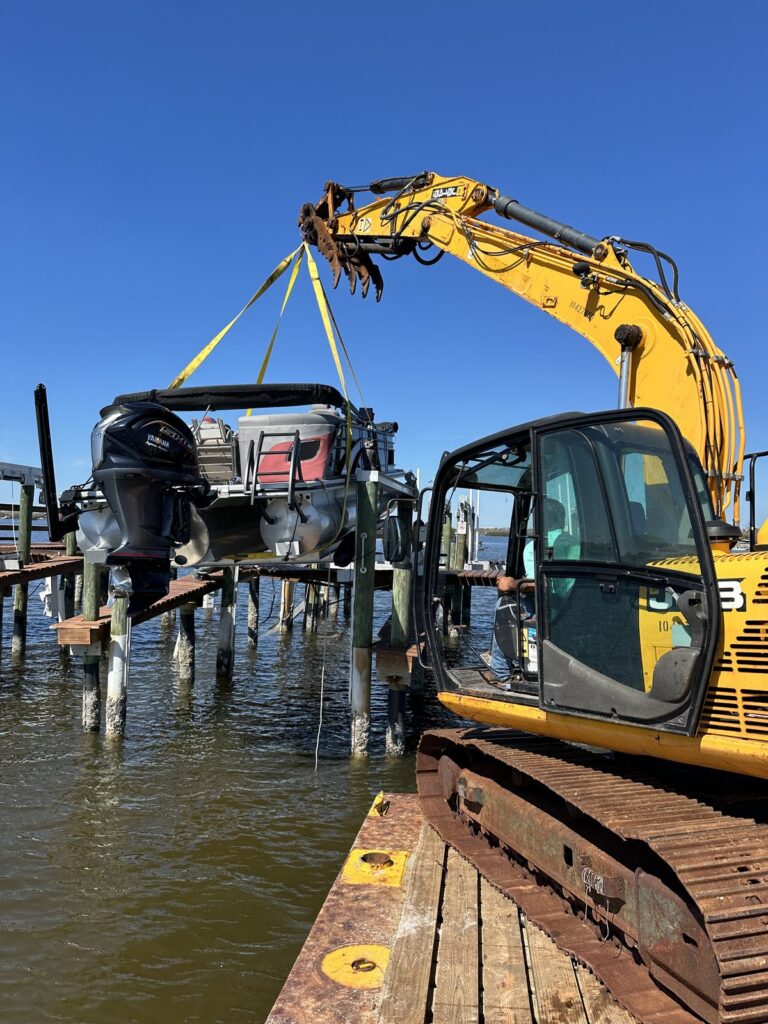 A tracked excavator on a steel work barge raises its boom and holds yellow lifting slings attached to a center console boat among weathered wood pilings. Barge based marine construction by Anderson Dock and Lift, Manatee County FL.