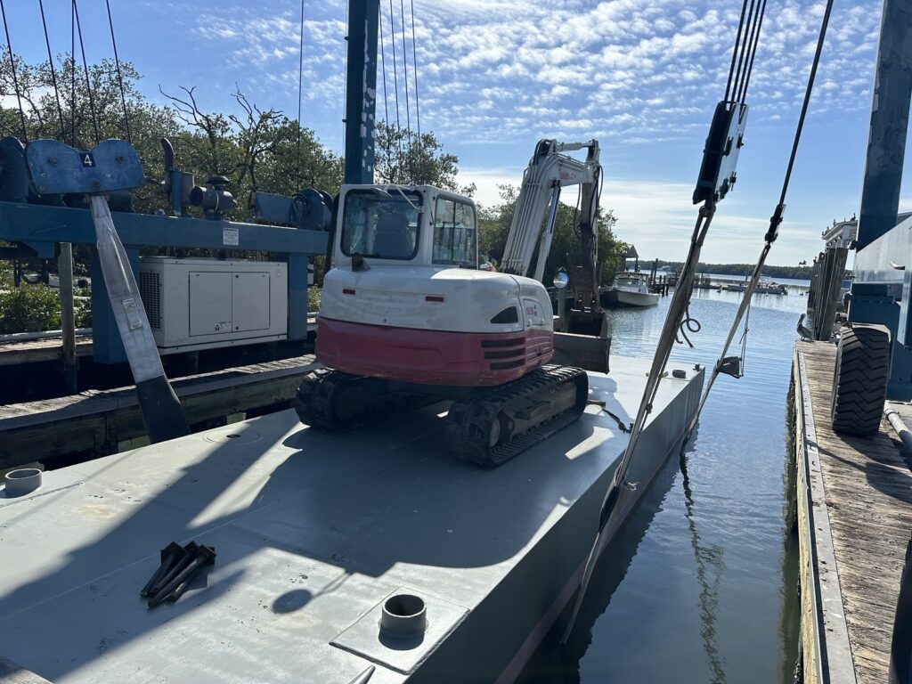 Tracked excavator parked on the deck of a steel work barge tied to a wooden dock, with a travel lift gantry and mangroves behind it. Marine construction equipment from Anderson Dock and Lift, Sarasota County FL.