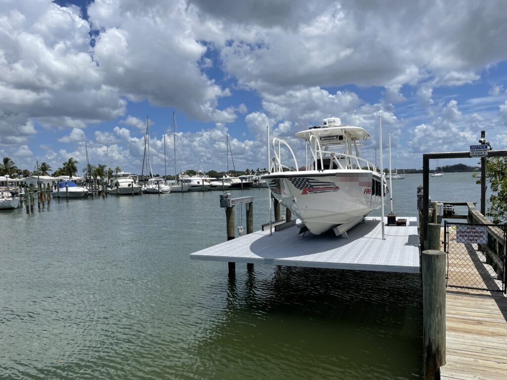 An aluminum boat lift platform holding a rescue boat at the end of a timber access dock, with a marina and moored sailboats across the basin. Commercial boat lift and dock construction by Anderson Dock and Lift, Sarasota County FL.