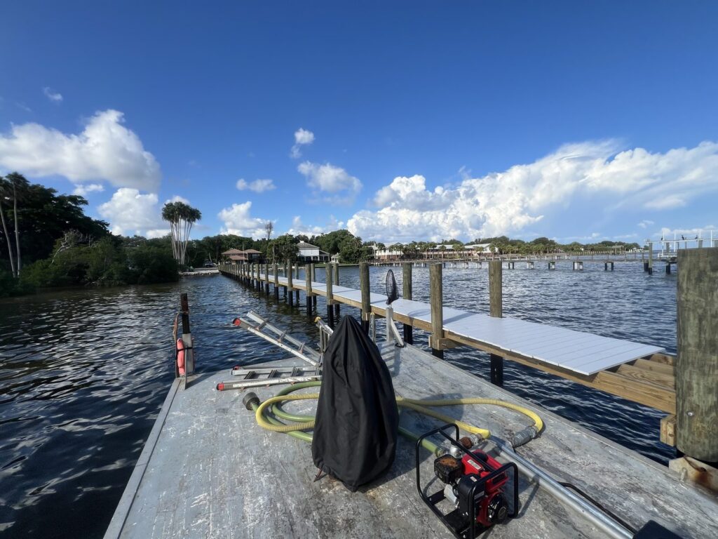 New pier on wood pilings with light gray deck panels set in place, seen from a work barge carrying a gas water pump, hoses, and ladders. Pier construction by Anderson Dock and Lift, Bradenton FL.