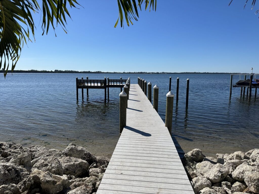 A dock walkway begins at a rock shoreline and runs out to an L shaped platform over the bay, framed by palm fronds. Dock and pier construction by Anderson Dock and Lift, Sarasota FL.
