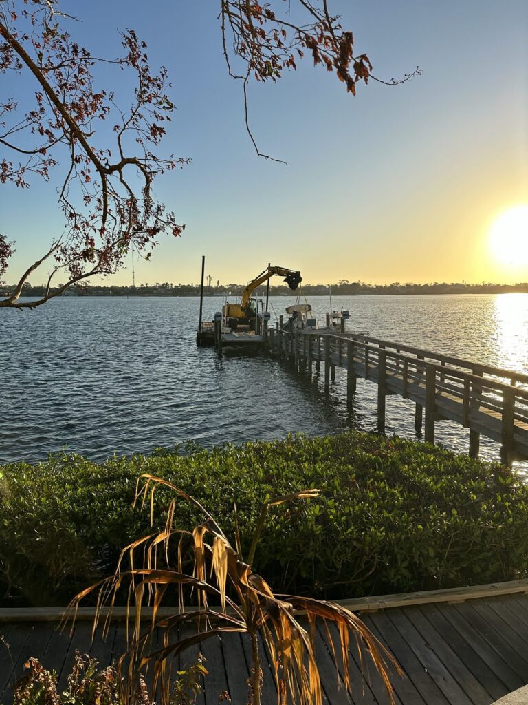 Excavator working from a barge to set new dock pilings at sunset. Dock piling installation by Anderson Dock and Lift, Sarasota FL.