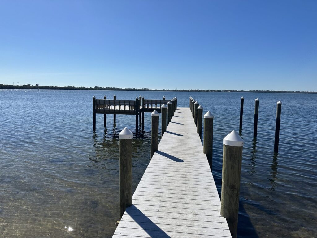 A dock walkway crosses shallow bay water to an L shaped platform with bench seating, carried on wood pilings with white conical caps. Dock and pier construction by Anderson Dock and Lift, Sarasota County FL.