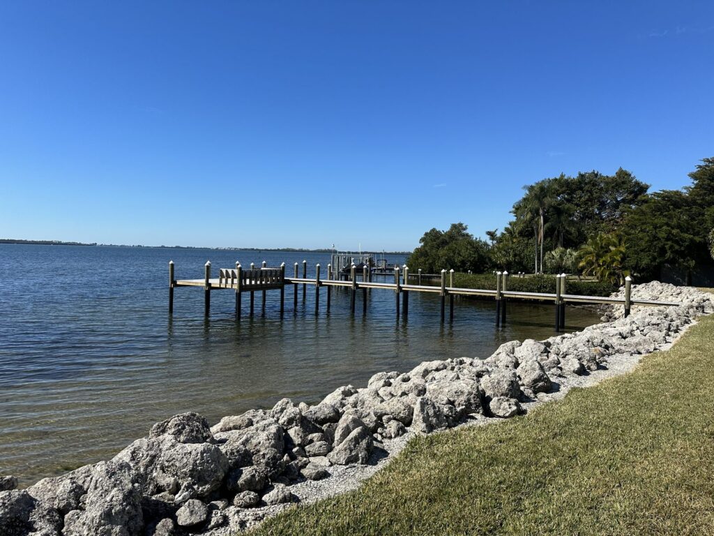 A rock bank armors a lawn shoreline where a dock runs out over the bay to an L shaped platform with bench seating. Dock and pier construction by Anderson Dock and Lift, Anna Maria Island FL.