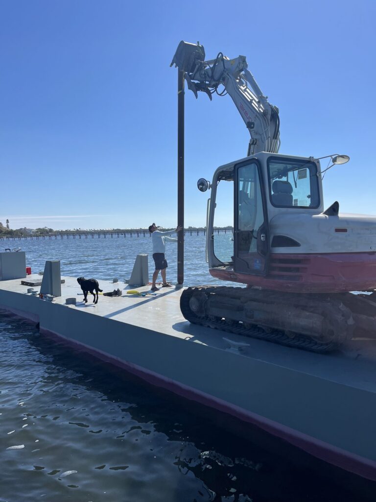 Tracked excavator and a long steel beam on the deck of a work barge crossing open water, with a worker standing at the rail. Marine construction crew at Anderson Dock and Lift, Manatee County FL.