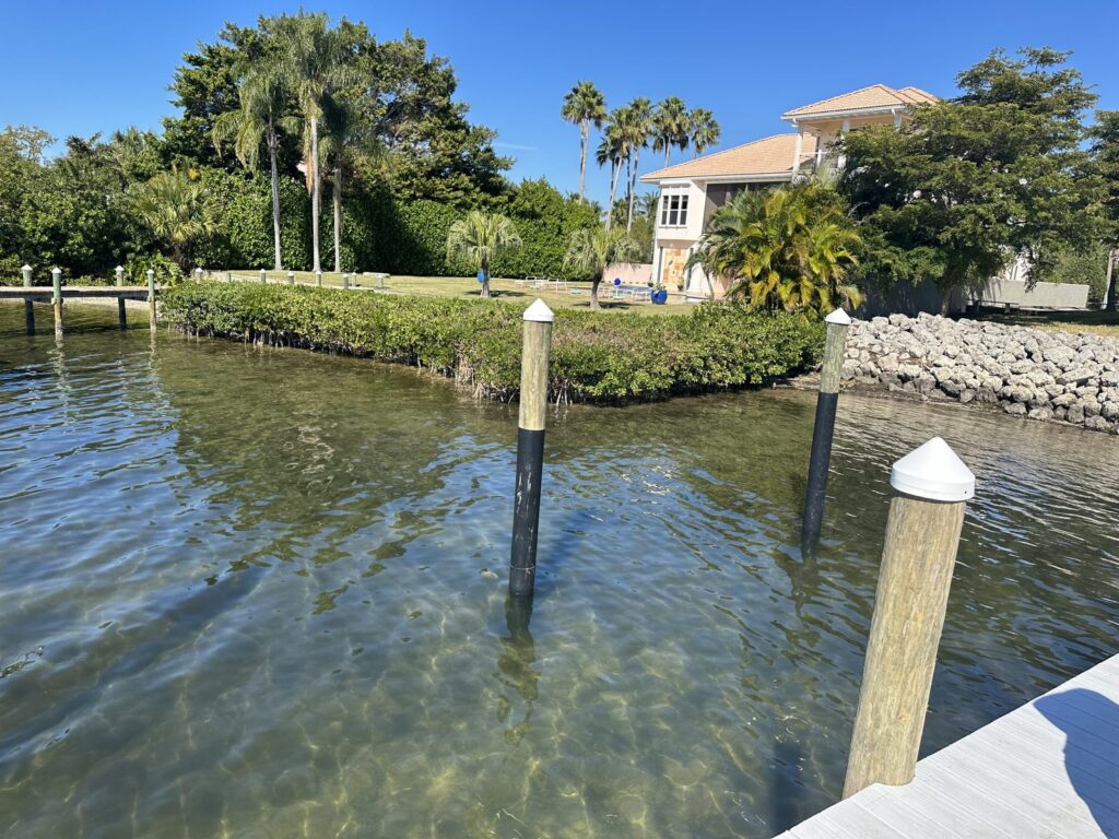 Wood pilings stand in shallow clear water with black wraps covering the sections at the waterline. Piling wrap installation by Anderson Dock and Lift, Bradenton FL.