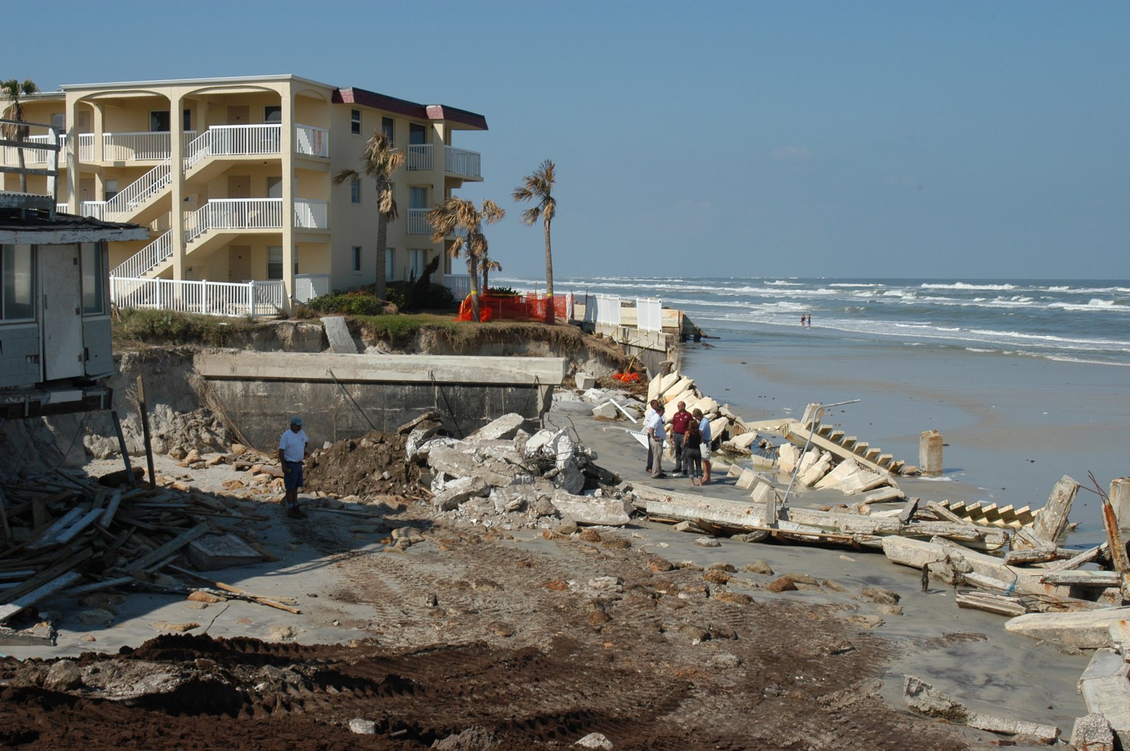 Concrete seawall still standing on a beach with the sand and fill washed out from behind it, the cap hanging over open air, broken wall sections on the sand below.