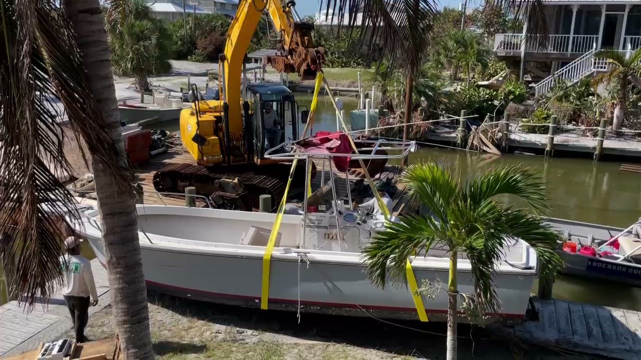 Excavator on a canal bank lifting a white center console boat in yellow straps after a storm, with an Anderson Dock and Lift workboat alongside.